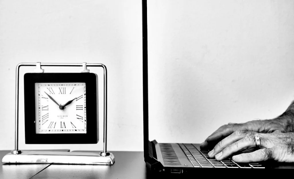 Reloj de escritorio analógico sobre una mesa junto a un portátil abierto mientras una persona escribe en el teclado, representando la gestión del tiempo y el trabajo frente al ordenador