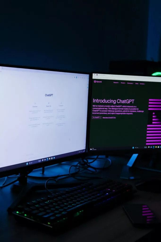 Dos monitores de ordenador encendidos en un escritorio oscuro, uno mostrando la página de ChatGPT y otro con el texto “Introducing ChatGPT”, junto a un teclado iluminado, representando el uso de herramientas de Inteligencia Artificial en el trabajo.