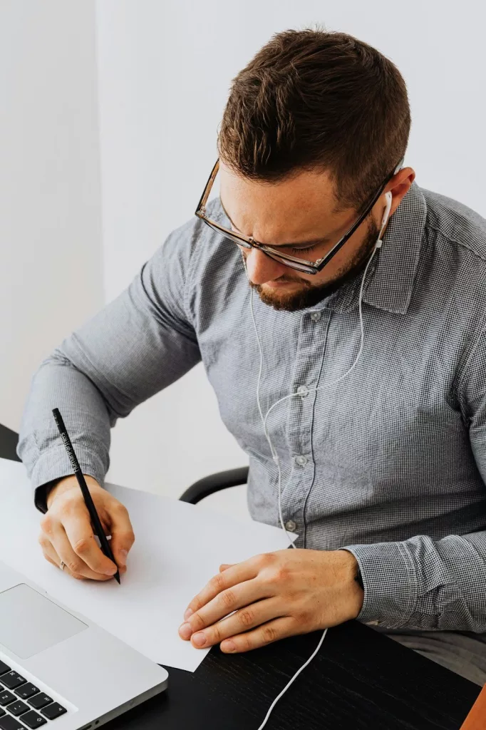 Hombre con gafas y auriculares escribiendo en una hoja de papel con un lápiz mientras trabaja en un escritorio junto a un ordenador portátil, representando el análisis y la planificación de tareas en un negocio.