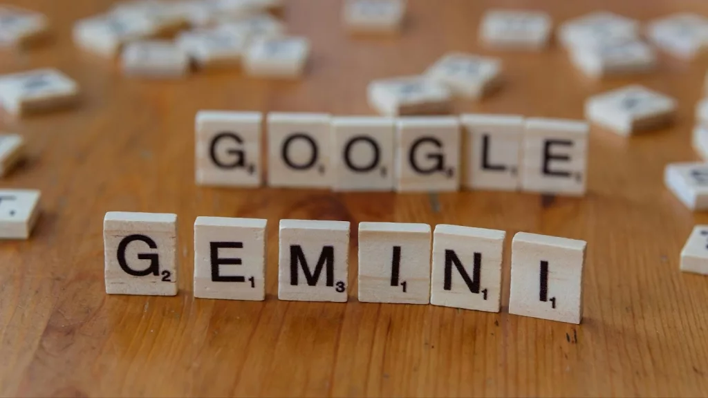 Letras de madera colocadas sobre una mesa formando la palabra “GEMINI” en primer plano y la palabra “GOOGLE” al fondo, ambas hechas con fichas tipo Scrabble, representando el modelo de inteligencia artificial Google Gemini en un entorno físico.