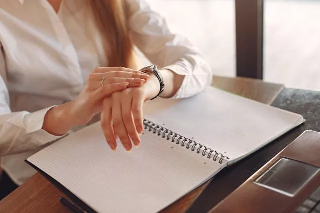 Persona mirando su reloj de pulsera mientras está sentada en un escritorio con cuaderno abierto y smartphone, reflejando control del tiempo en el entorno laboral
