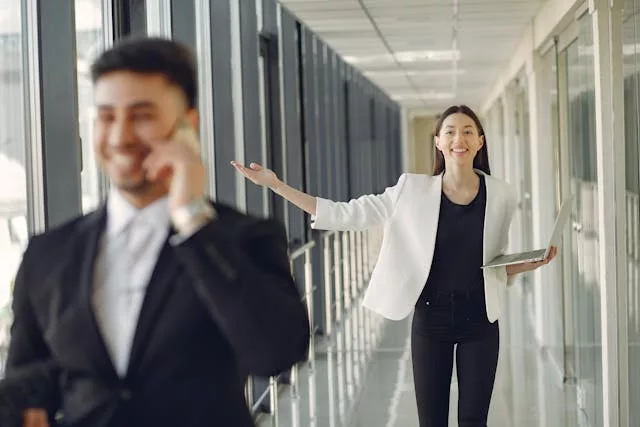 Mujer con blazer blanco sosteniendo una tablet señalando en un pasillo de oficina mientras un hombre habla por teléfono en primer plano desenfocado