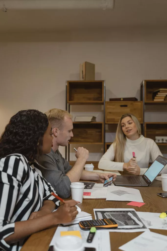 Tres personas reunidas alrededor de una mesa de madera en una oficina, con un portátil abierto, vasos de café, papeles, notas adhesivas, una calculadora, un móvil y rotuladores sobre la mesa, mientras una mujer rubia habla y las otras dos personas escuchan y toman notas.