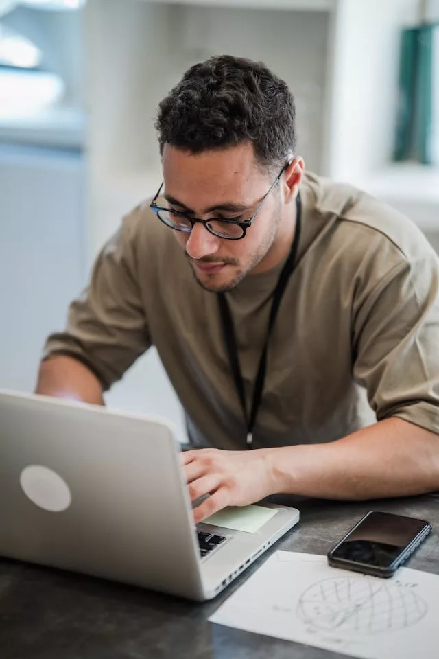 Un hombre joven con gafas y camiseta beige busca información en su ordenador portátil para aprender IA desde cero. En primer plano se aprecia su mano sobre el teclado, un teléfono móvil a la derecha y un papel con diagramas dibujados a mano sobre una mesa gris oscura.