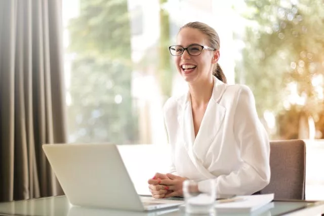 Mujer adulta de mediana edad, bien vestida, sonriendo mientras trabaja con un portátil en una oficina luminosa con ventanas al fondo.