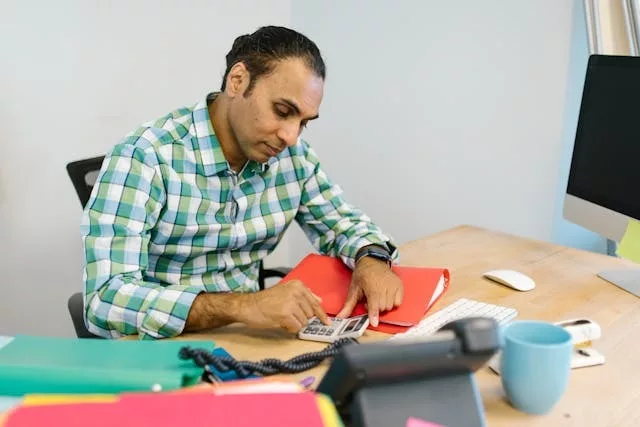 Hombre adulto concentrado usando una calculadora en su escritorio de oficina, con documentos, carpetas y una taza alrededor, trabajando en cálculos financieros.